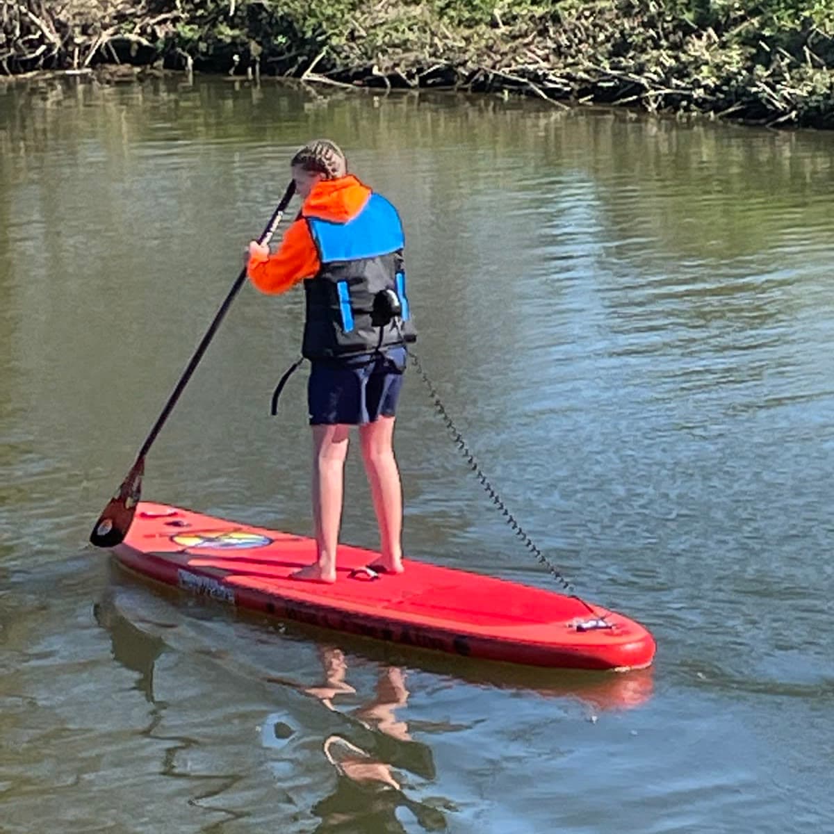 A young paddler in a red life jacket standing on a NewWave board on the Stour
